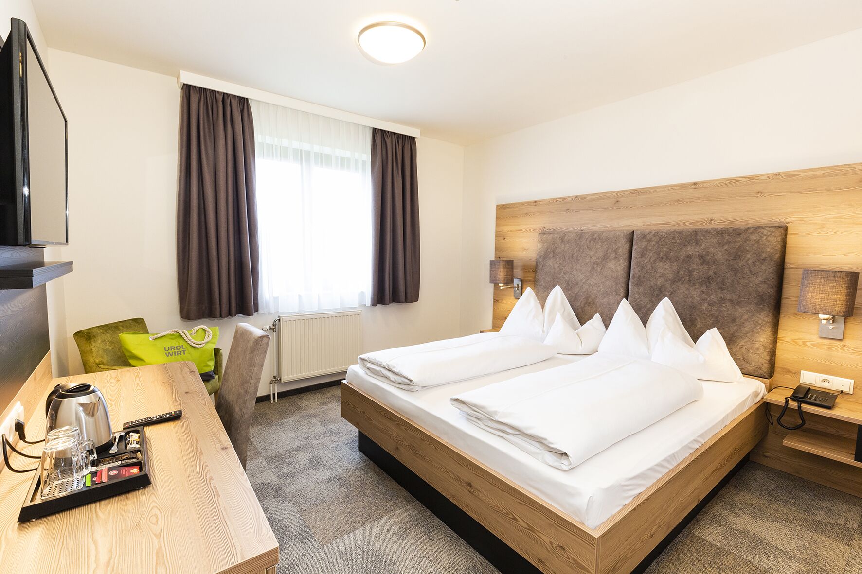 A hotel room with two beds, a wooden headboard, a desk with a kettle, glasses, and a remote control.