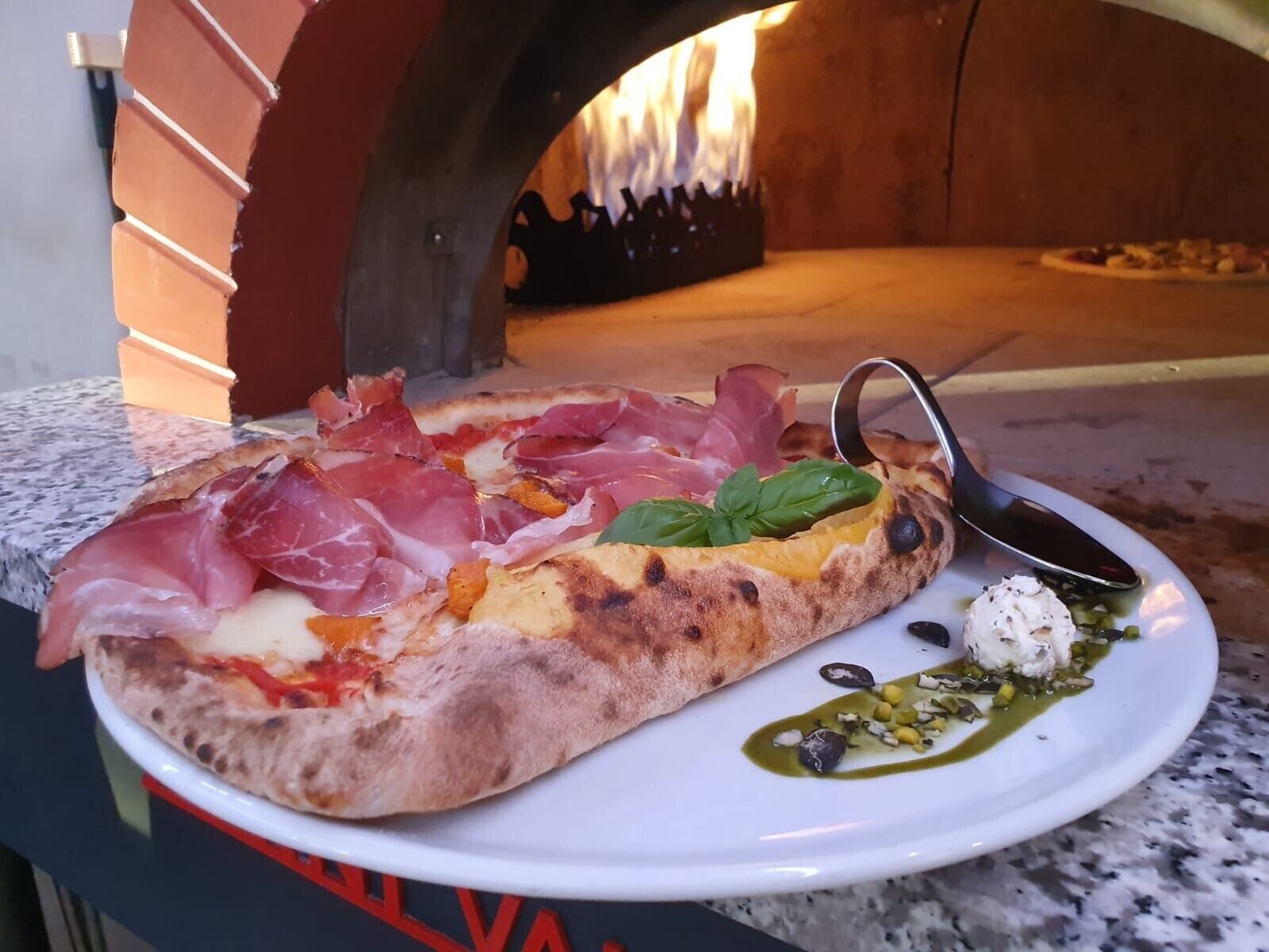 A pizza with prosciutto and basil on a white plate, placed on a granite countertop.