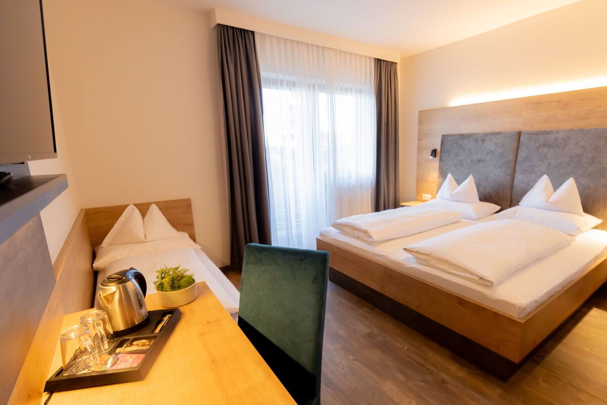 Une chambre d’hôtel avec un grand lit, un bureau avec une bouilloire et une petite plante, et une fenêtre avec des rideaux.