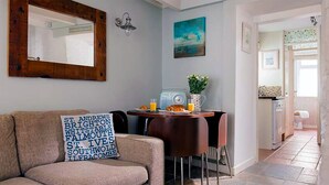 Un salon chaleureux avec un canapé, une table basse et un coin repas comprenant une table et des chaises. On aperçoit une cuisine au fond de la pièce.