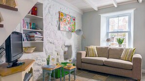 Un salon avec un mur en pierre blanche, un canapé beige, un téléviseur, une bibliothèque et une fenêtre ornée de plantes.