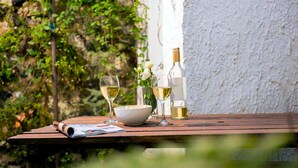Une table en bois, sur laquelle sont disposés un saladier, des verres à vin et des bouteilles, est placée devant un mur blanc, avec de la verdure en arrière-plan.