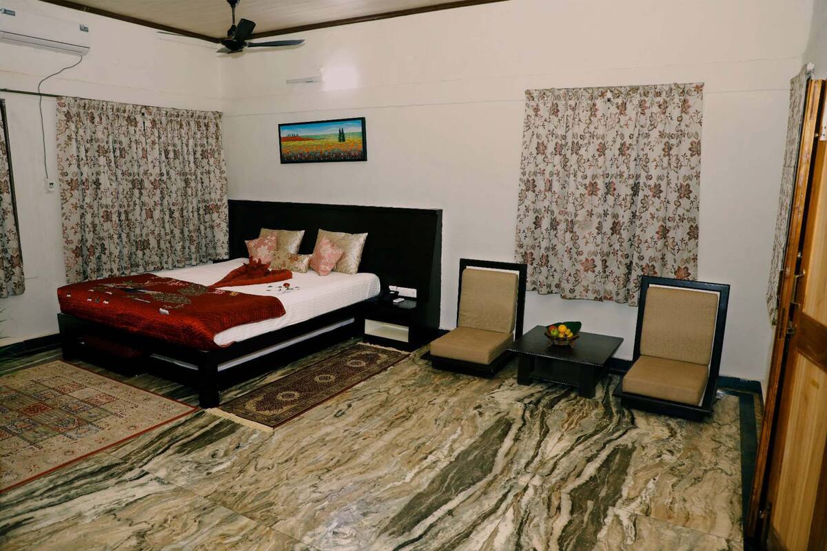 Een slaapkamer met een bed, twee stoelen, een klein tafeltje met een schaal, een plafondventilator en een schilderij aan de muur.