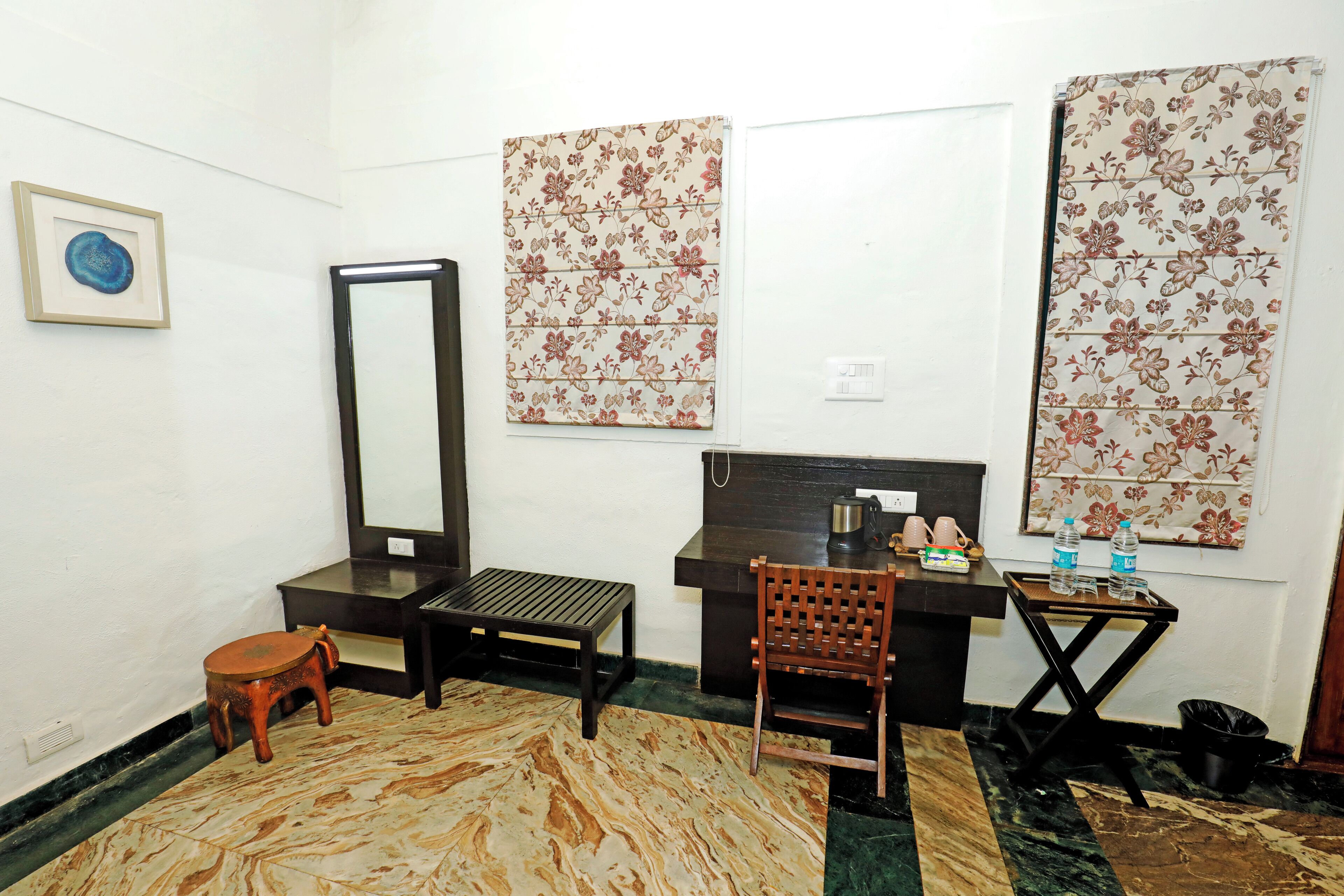 A small, furnished room with a desk, chair, and a mirror. There are two windows with floral curtains, a small stool, and a trash bin.