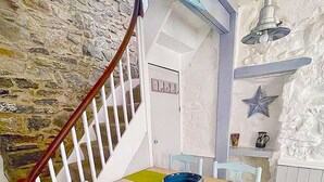 A dining area with a wooden table, blue chairs, a yellow tablecloth, a stone wall, a white door, and a staircase with a red railing. - St Ives town cottage - family & pets - sleeps 4, Holiday Cottages St Ives (St Ives)