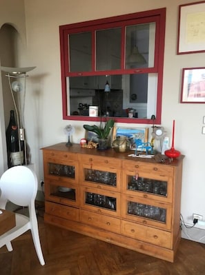 Een houten kast met glazen deuren, een raam met rode omlijsting erboven en een witte stoel links.
