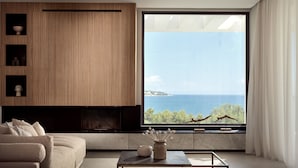A modern living room with a large window overlooking the sea, a built-in wooden panel wall, and a minimalist coffee table. - Omnia Villa (Zakynthos)