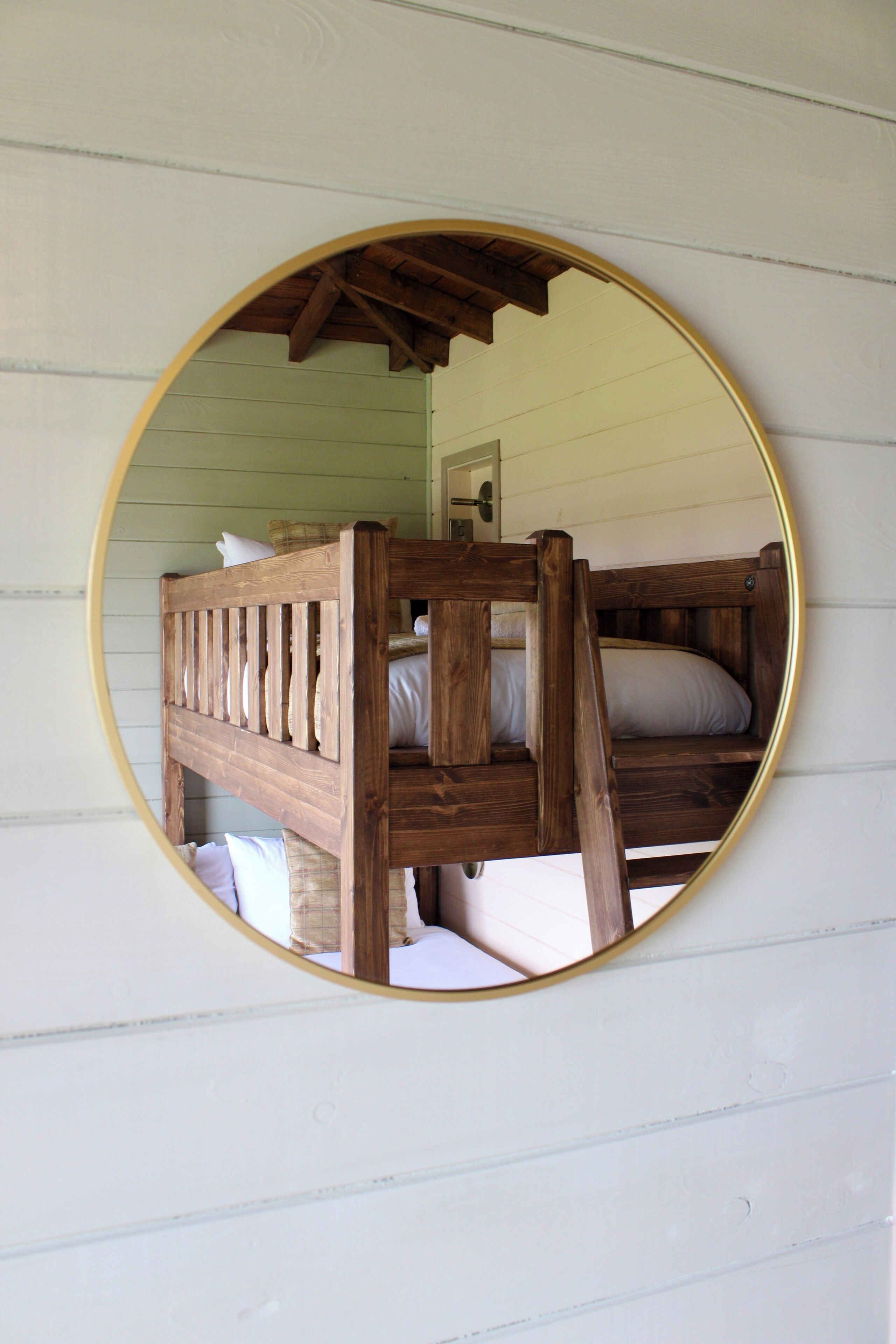 Una cama litera con estructura de madera y ropa de cama blanca, vista a través de un espejo circular.