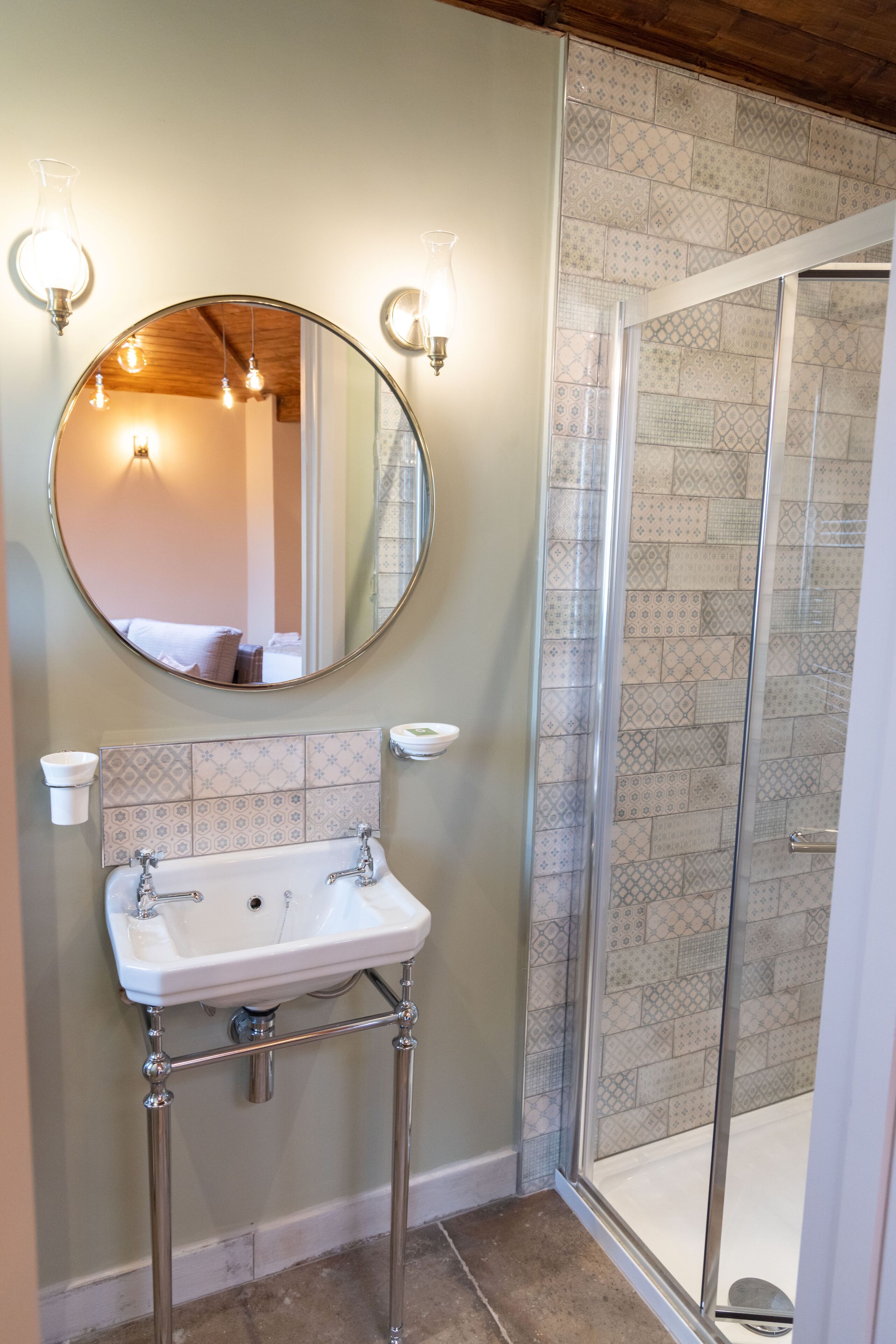 A bathroom with a round mirror, two wall-mounted lights, a white sink with chrome fixtures, and a patterned tile backsplash.