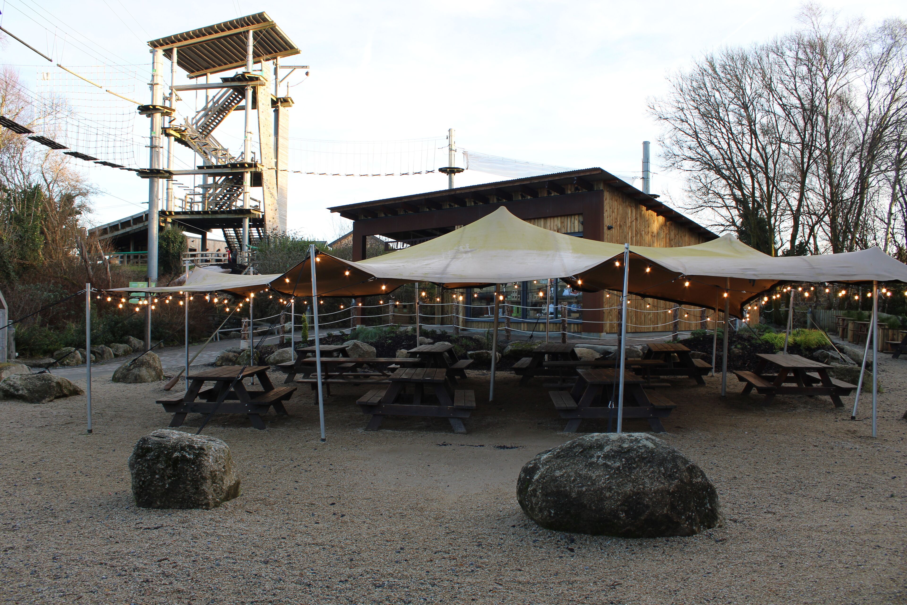 An outdoor dining area with picnic tables and umbrellas, a wooden building, and a rope bridge.