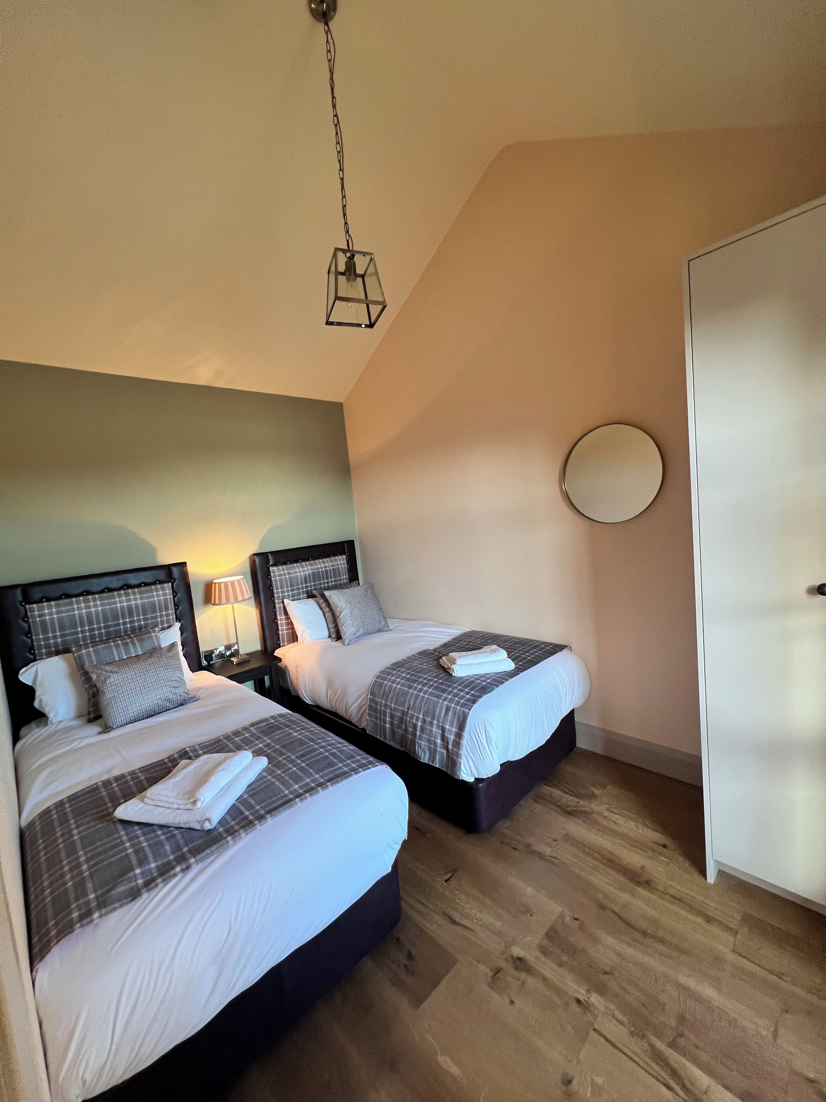A bedroom with two beds, a bedside table with a lamp, a round mirror, and a wardrobe.