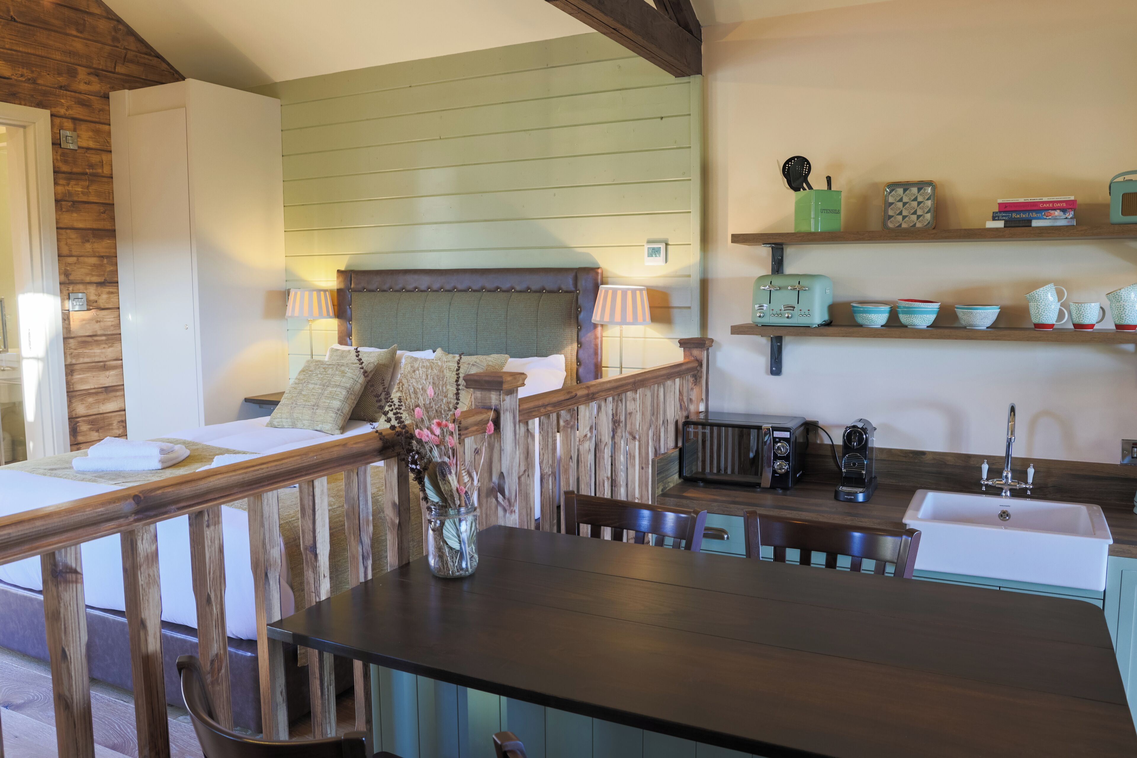 A cozy room with a bed, a wooden table with a vase, a kitchen area with a sink and retro appliances, and a shelf with decorative items.