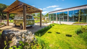 A green vintage van parked under a wooden pergola, a modern glass building, and a well-maintained grassy area with rocks.