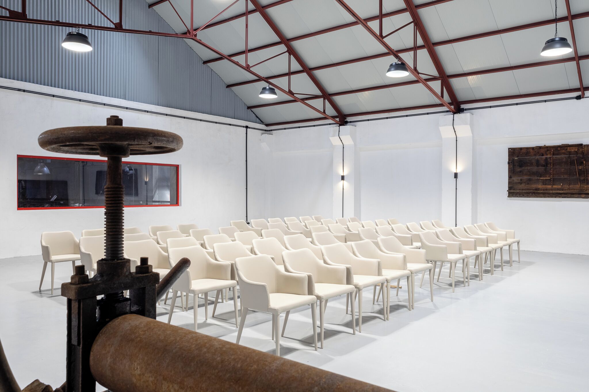 Ein geräumiger Raum mit Reihen weißer Stühle, einem großen Tisch im Industriedesign und einer hohen Decke mit sichtbaren Balken.