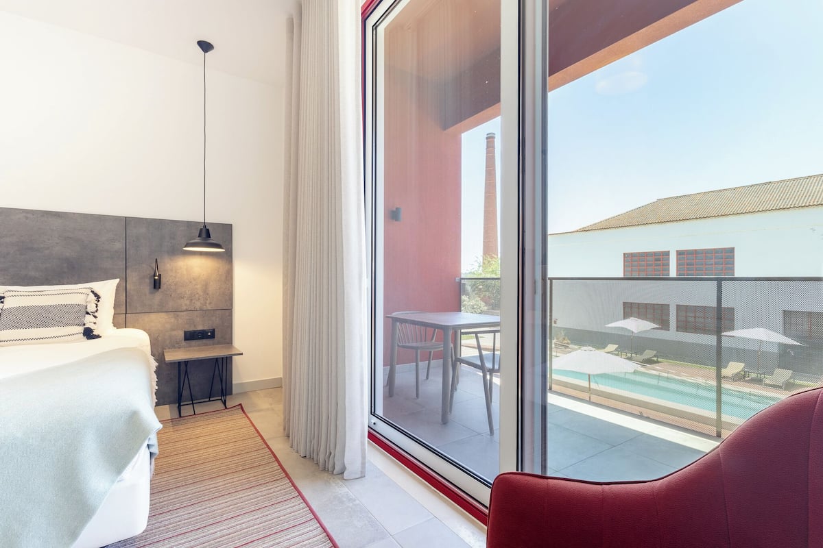 Moderni hotellihuone, jossa on suuri liukuovi, joka johtaa parvekkeelle, valkoisella päätyosalla varustettu sänky, pieni pöytä ja punainen tuoli.