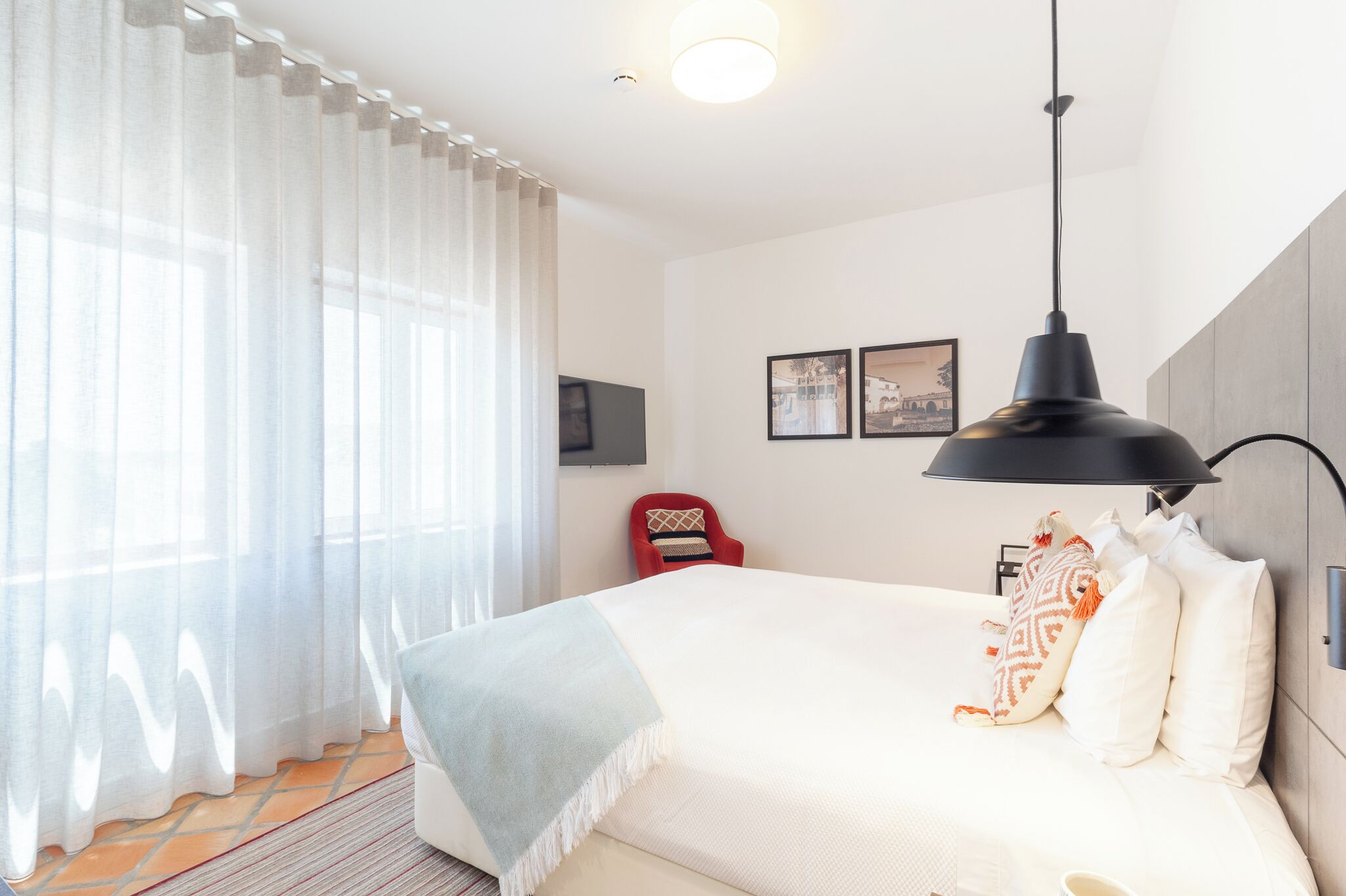 A hotel room with a bed, a red chair, a black pendant lamp, and two framed pictures on the wall.