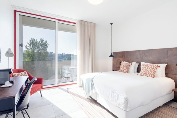 A modern hotel room with a large bed, a desk with a chair, a balcony with a view, and a sliding glass door. - Barrosinha Hotel (Alcacer do Sal)