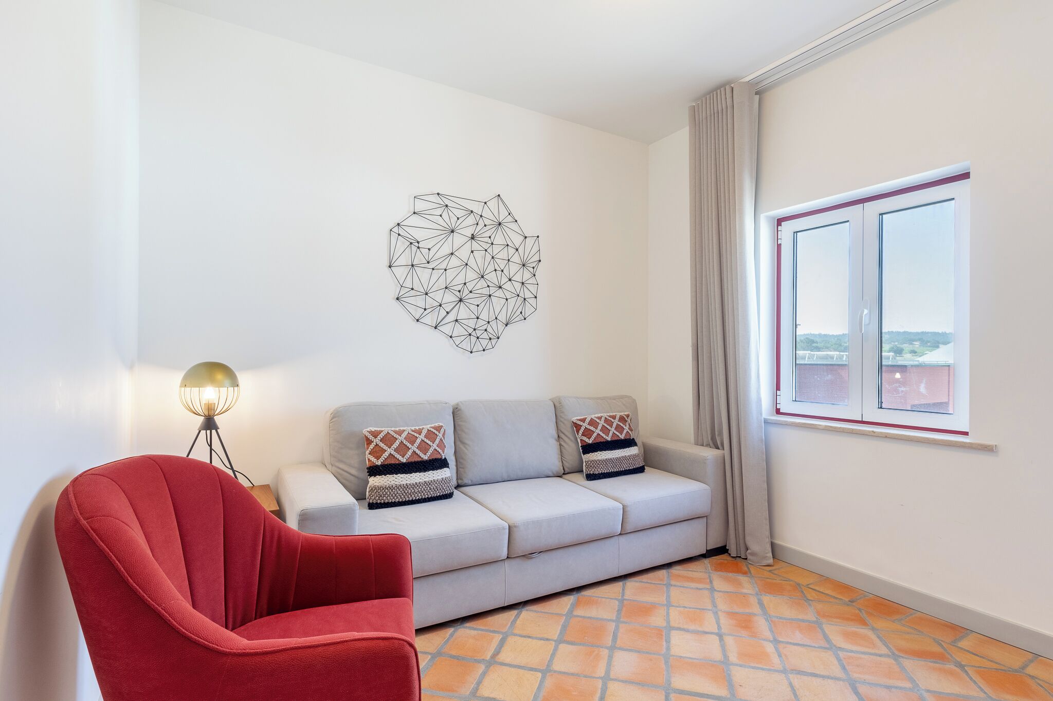 A modern living room with a red armchair, a grey sofa with patterned cushions, a geometric wall art piece, and a window with a view.
