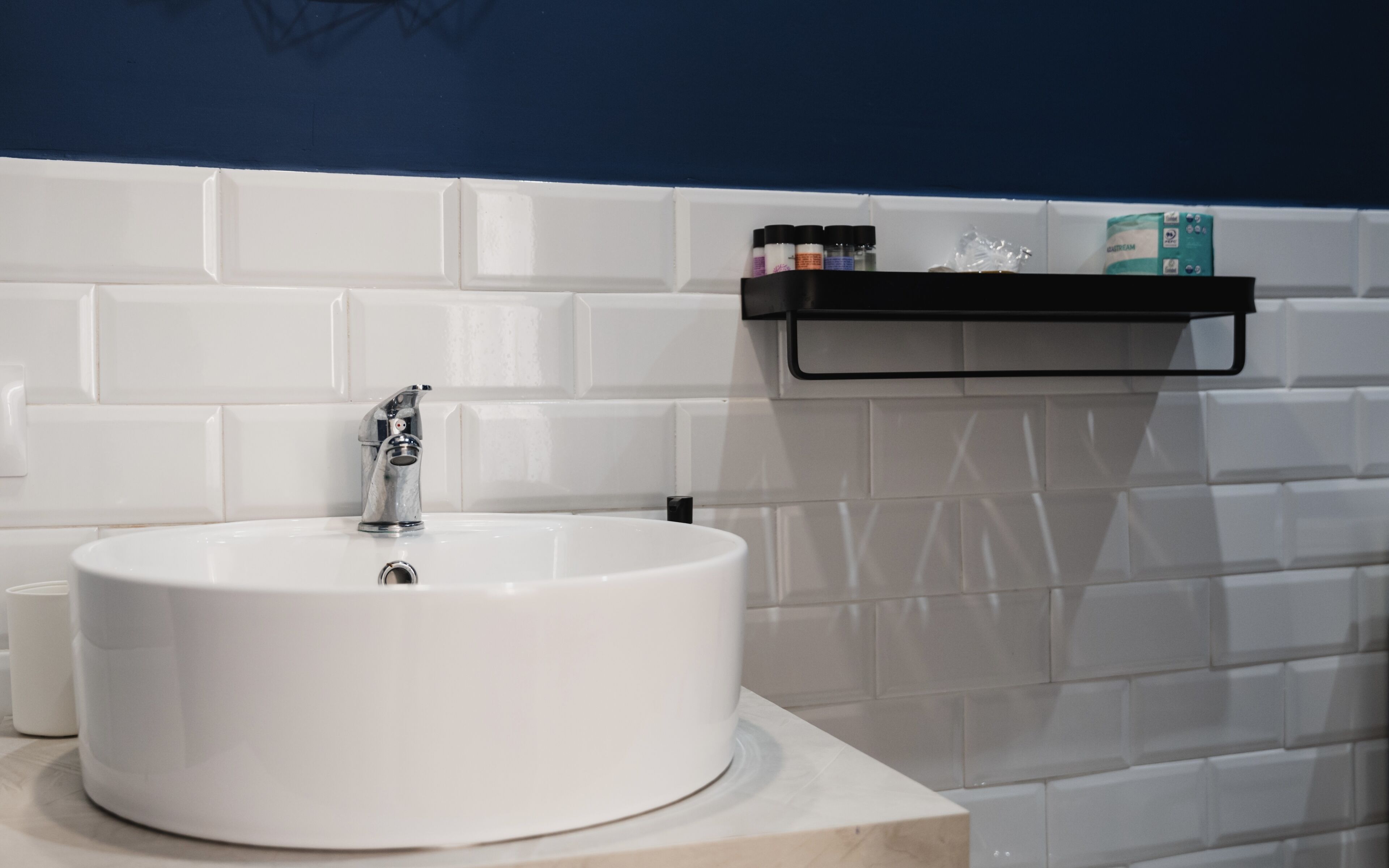 Un lavamanos blanco con un grifo moderno, un jabonera negra y una pared con azulejos blancos.