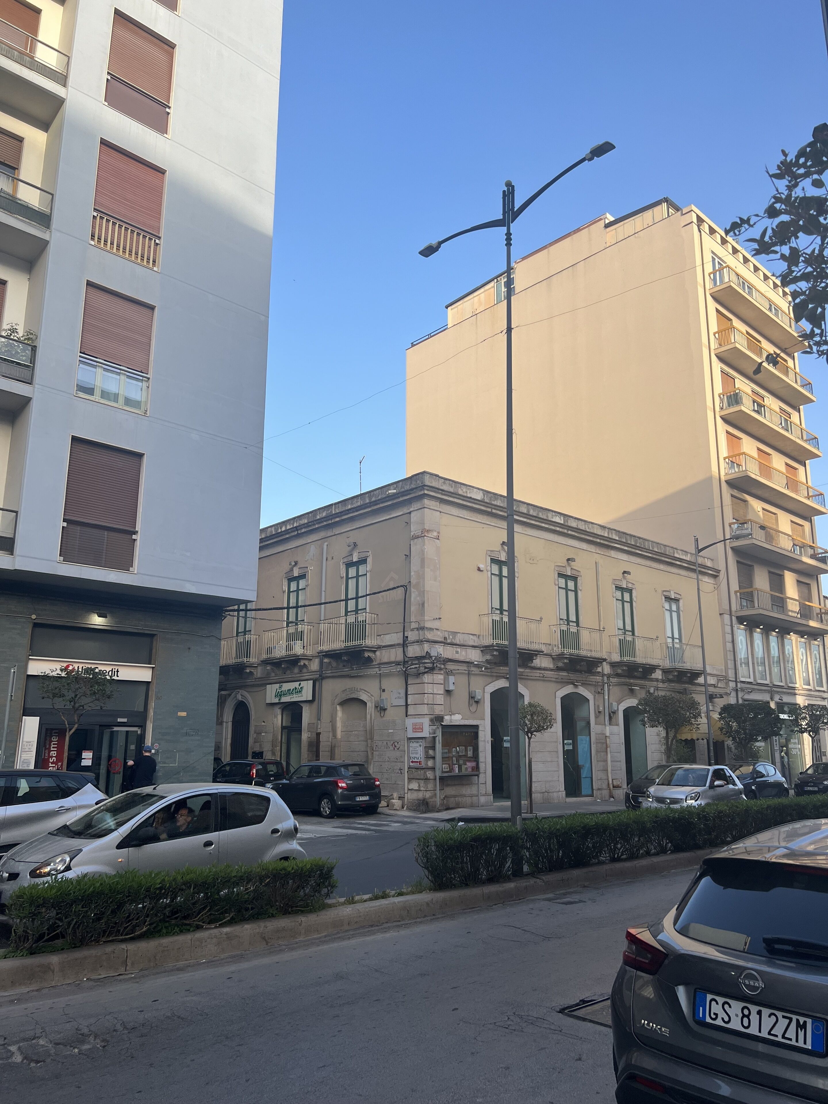 Un edificio de varios pisos con fachada color crema, una calle al frente y automóviles estacionados.