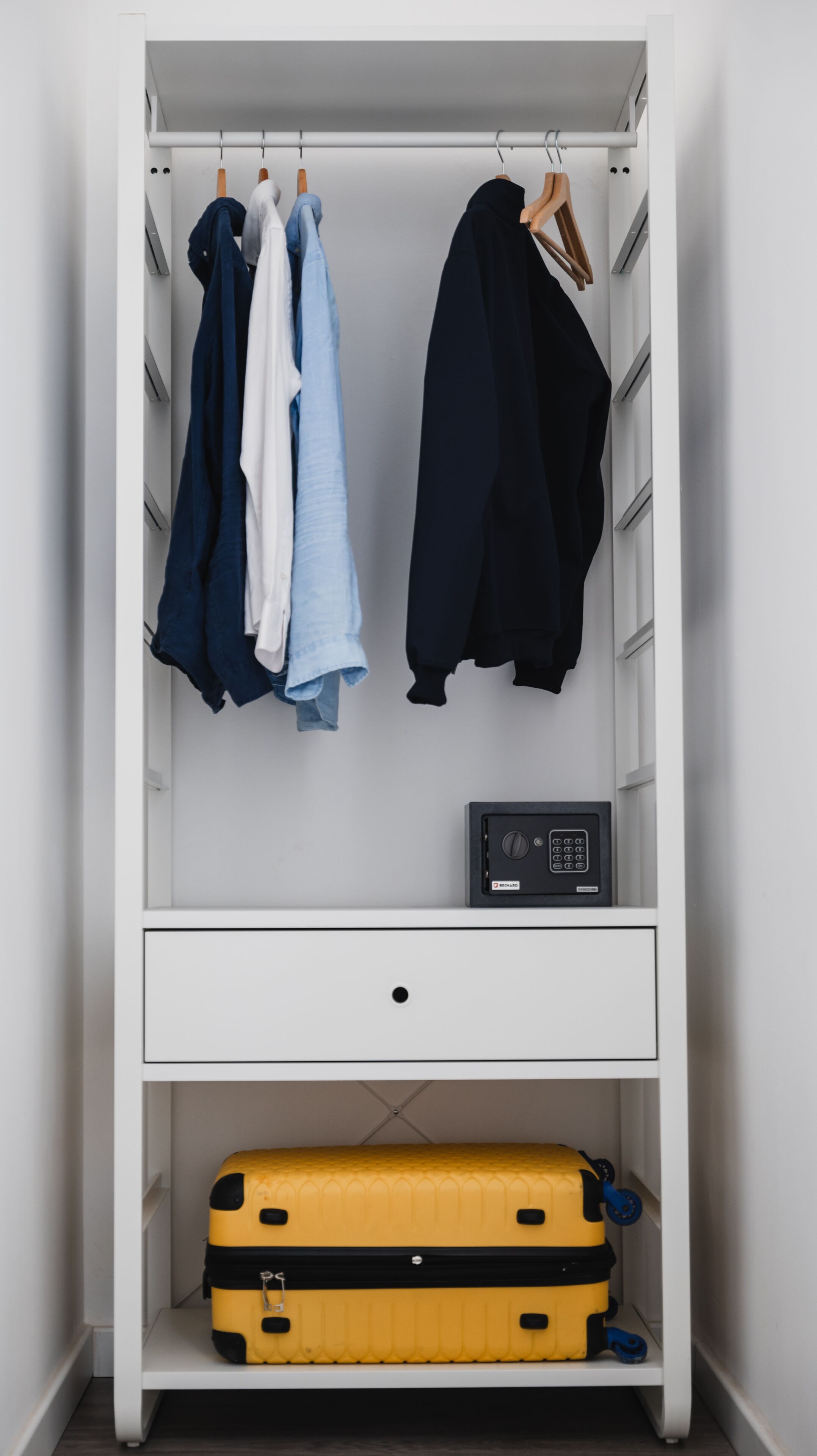 A white wardrobe with hanging clothes, a black electronic device, and a yellow suitcase on the bottom shelf.