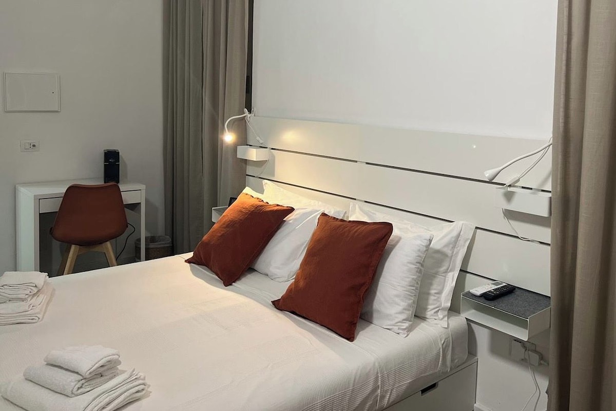A neatly made bed with white and brown pillows, a bedside table with a remote control, and a chair with a desk.
