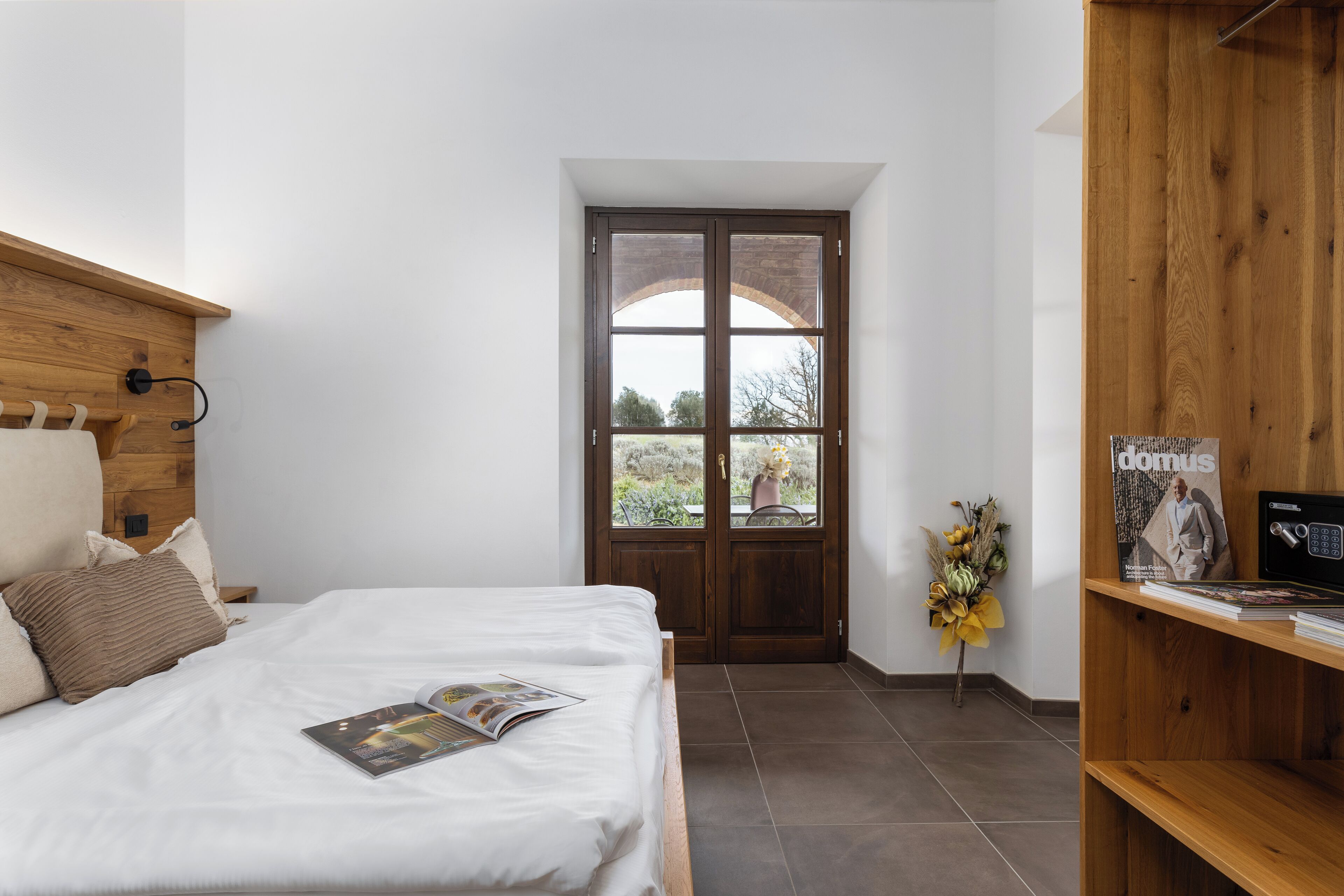 Un dormitorio con cabecera de madera, cama con ropa blanca, una revista sobre la cama, estantería de madera y una puerta que da a un balcón con vistas a árboles y una estructura de ladrillo.