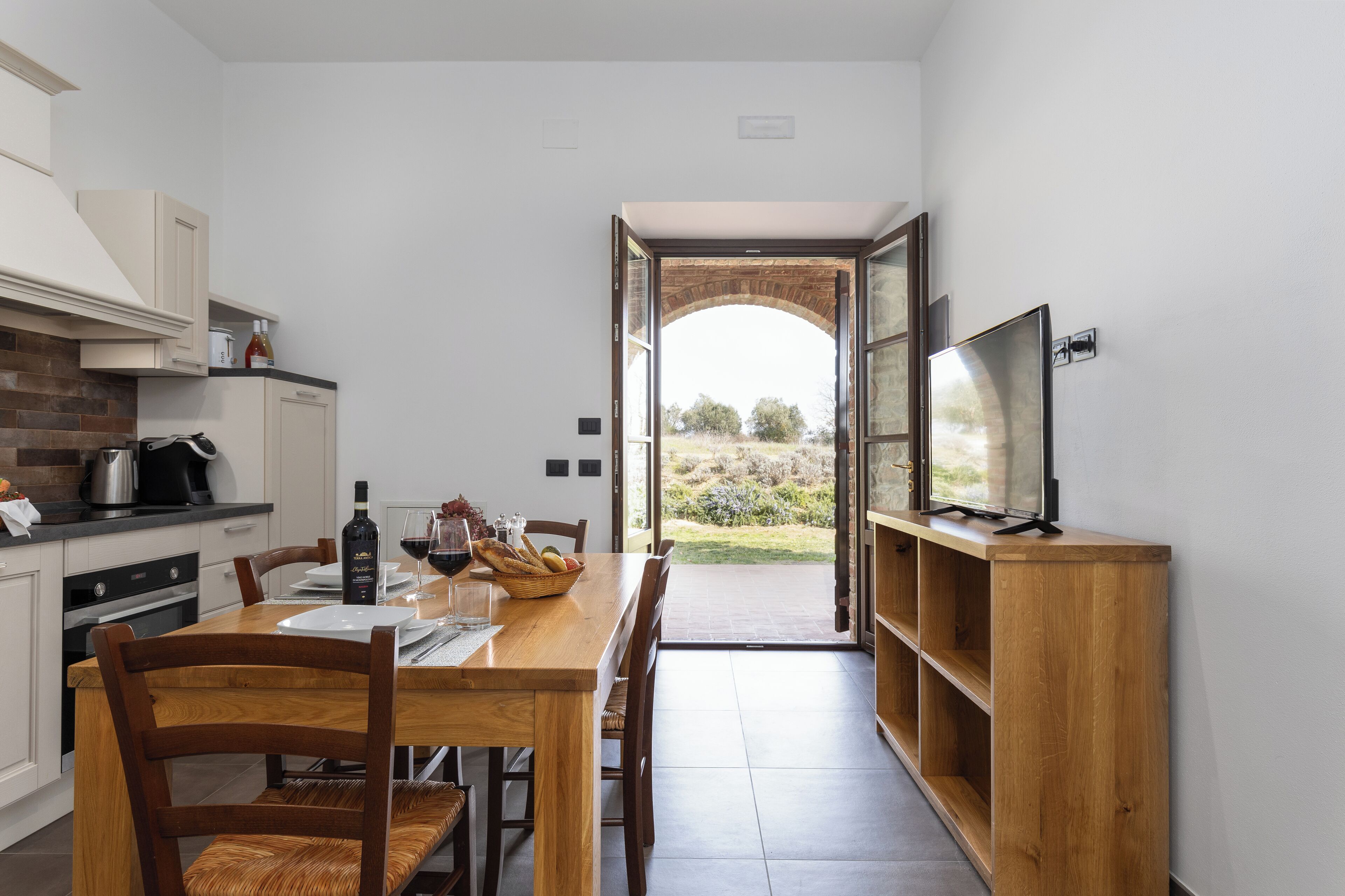 A modern kitchen with a dining area, a TV on a wooden stand, and a view of the outdoors through an open archway.