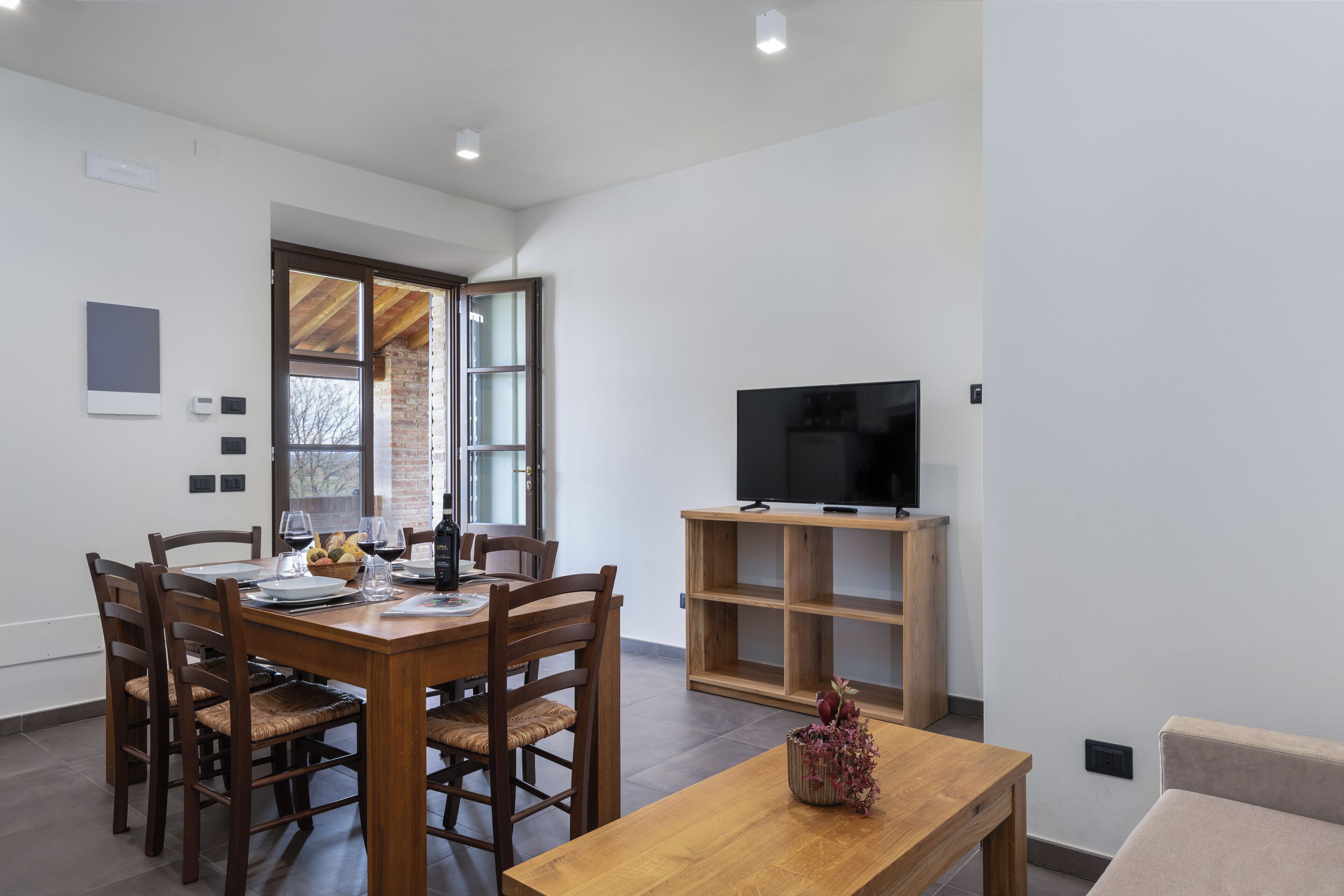 A modern living room with a wooden dining table, chairs, a TV on a wooden stand, and a small table with a vase.
