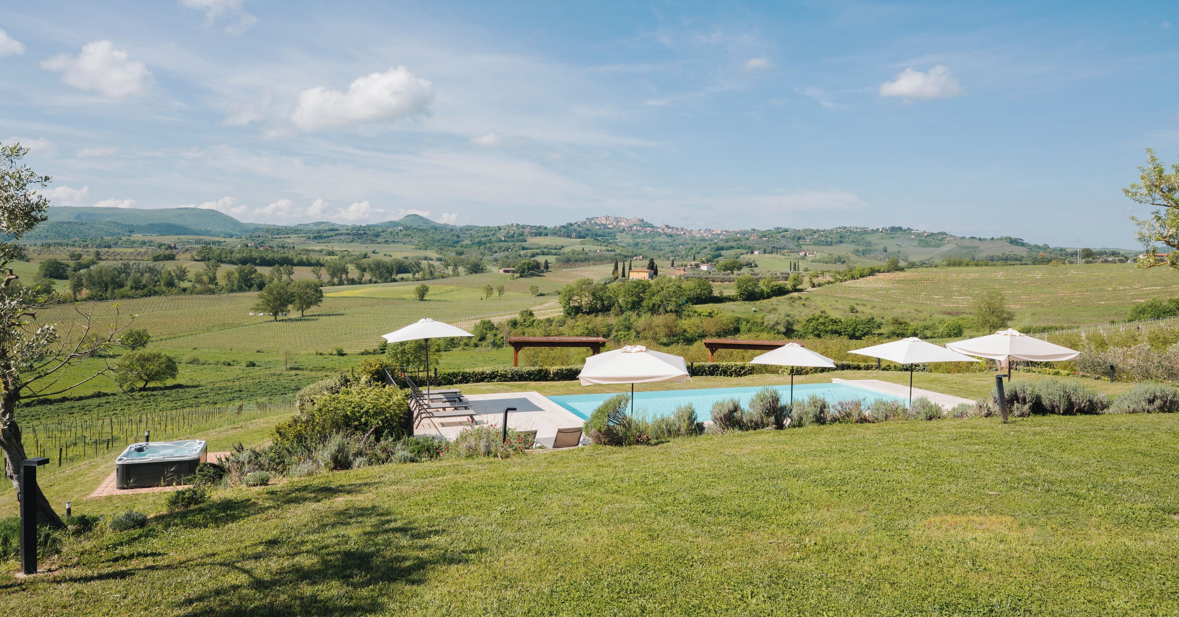A pool area with umbrellas, a hot tub, and a view of a vineyard and distant hills.
