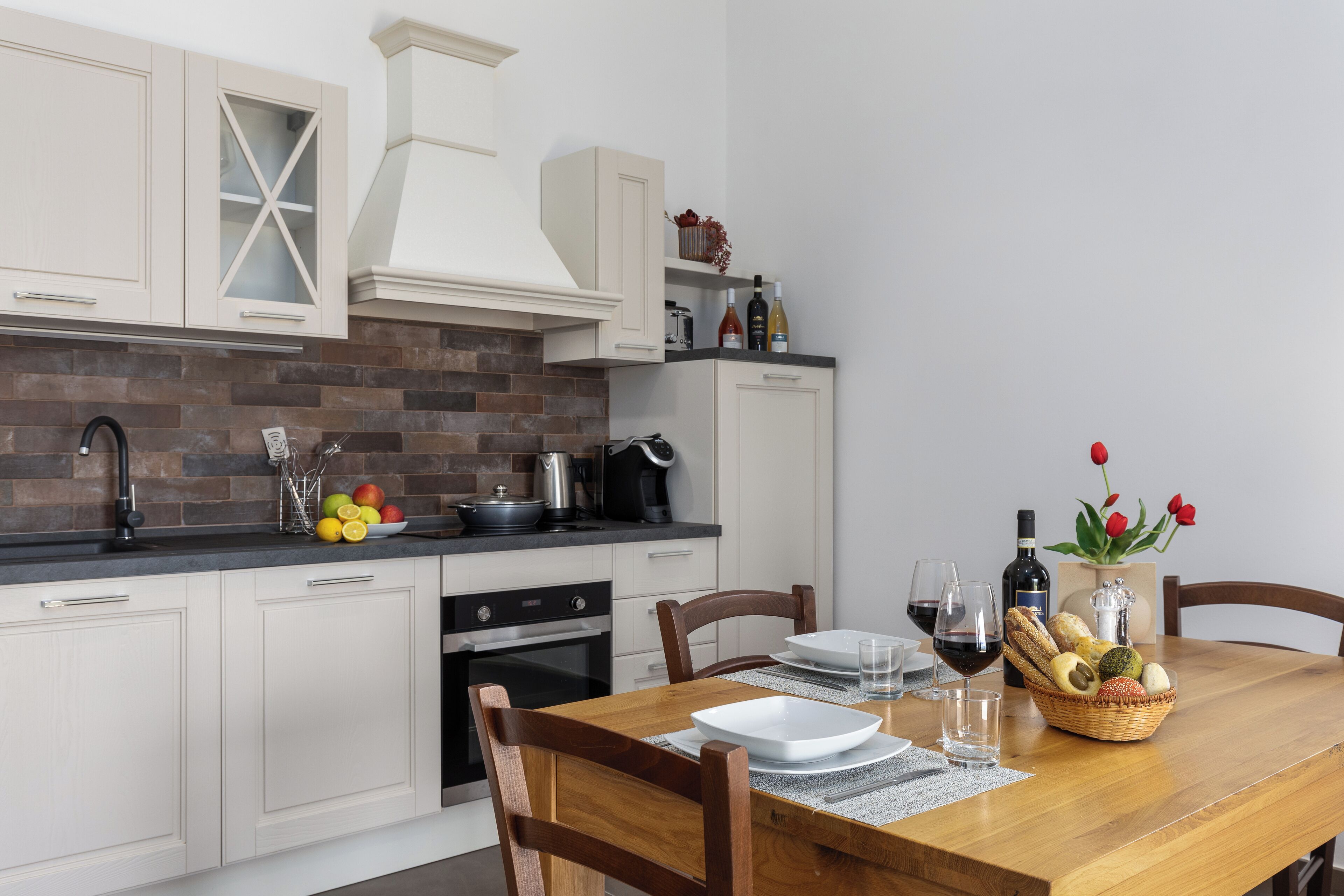 A modern kitchen with white cabinetry, a brick backsplash, and a dining table set with plates, glasses, and a fruit basket.