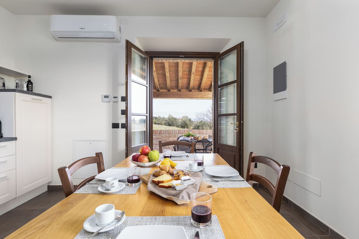 A dining area with a wooden table set for breakfast, including fruits, bread, and beverages.