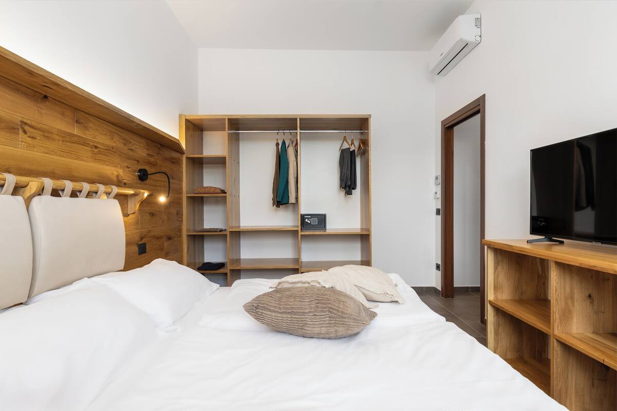 A bedroom with a wooden headboard, a bed with white bedding, a wooden wardrobe with hanging clothes, a television on a wooden stand, and a small bedside table.