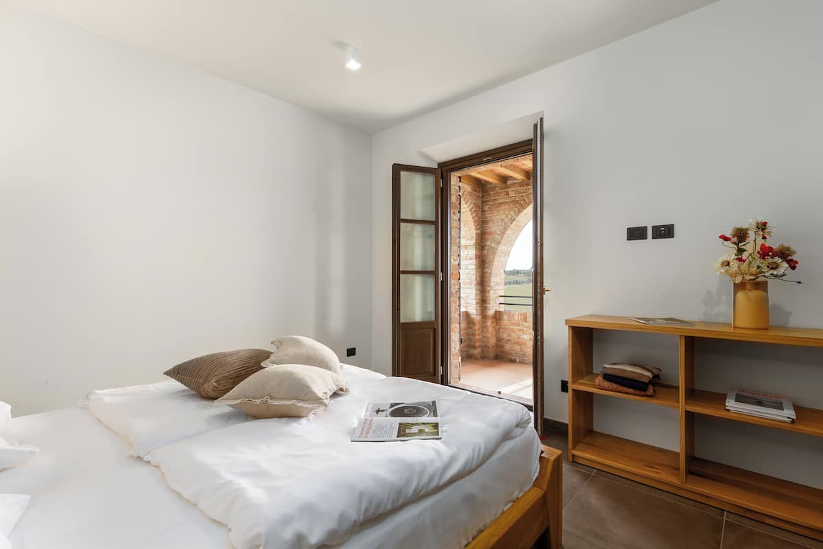 A bedroom with a bed, a wooden shelf, and a view through an open door to a brick structure.