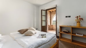 Quarto com cama, prateleira de madeira e vista através de uma porta aberta para uma construção de tijolos.