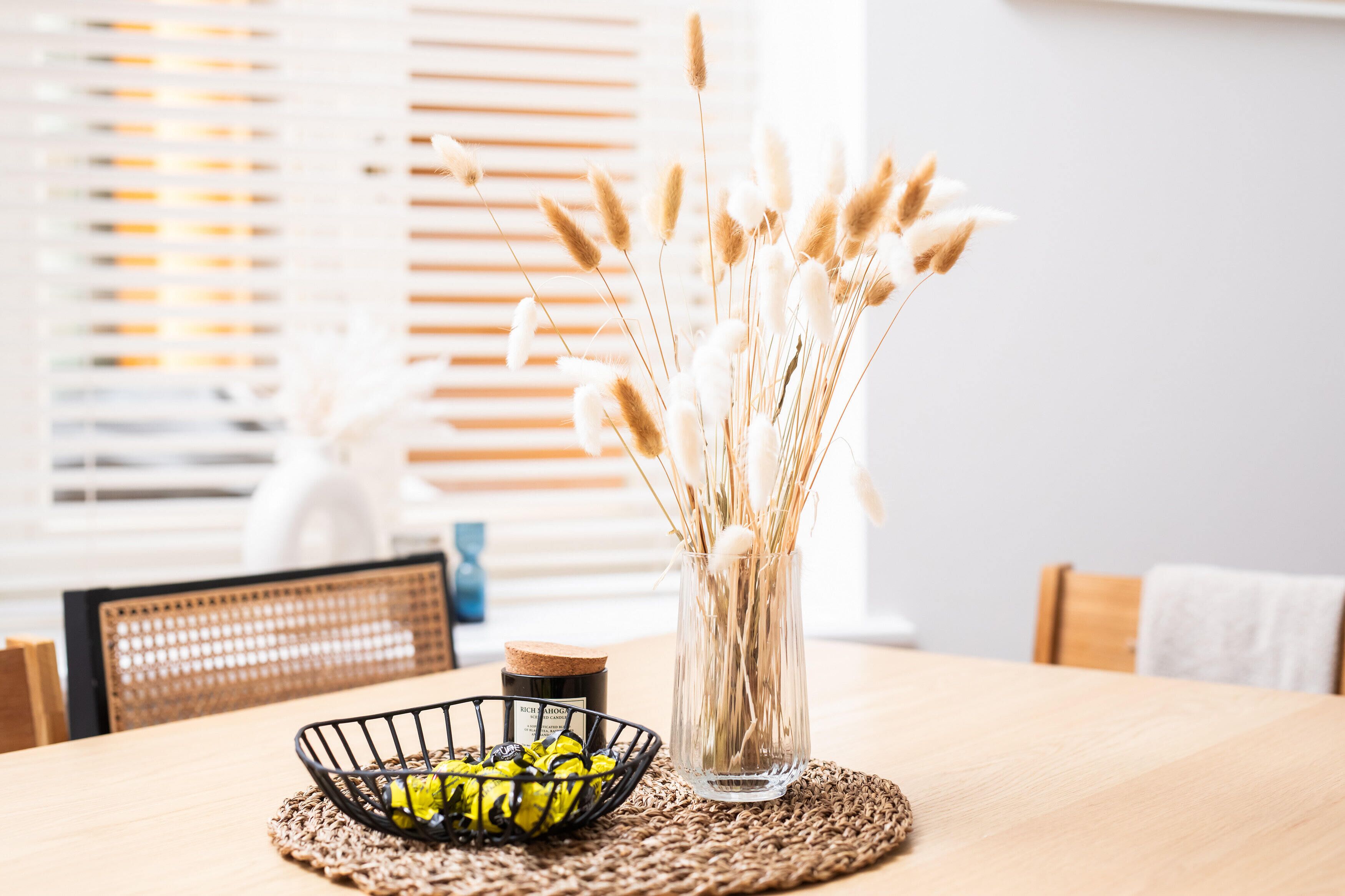 Une table à manger ornée d’un vase en verre transparent contenant des herbes des pampas séchées, d’un panier en fil de fer noir avec des fleurs jaunes et d’un petit bocal.