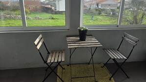 Et værelse med udsigt over et grønt landskab gennem store vinduer, et lille bord med en potteplante og to træstole.