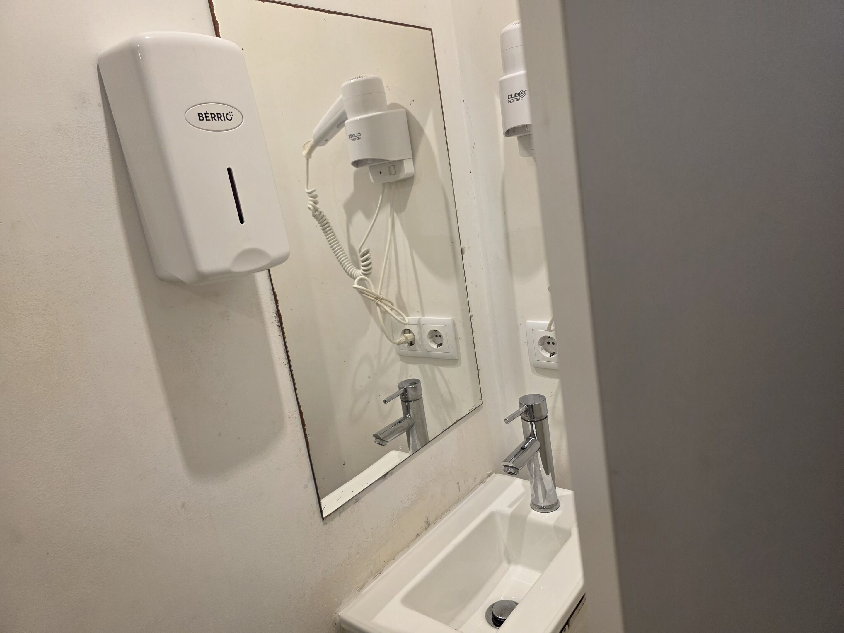 Une salle de bain avec un lavabo blanc, un miroir et un sèche-cheveux fixé au mur.