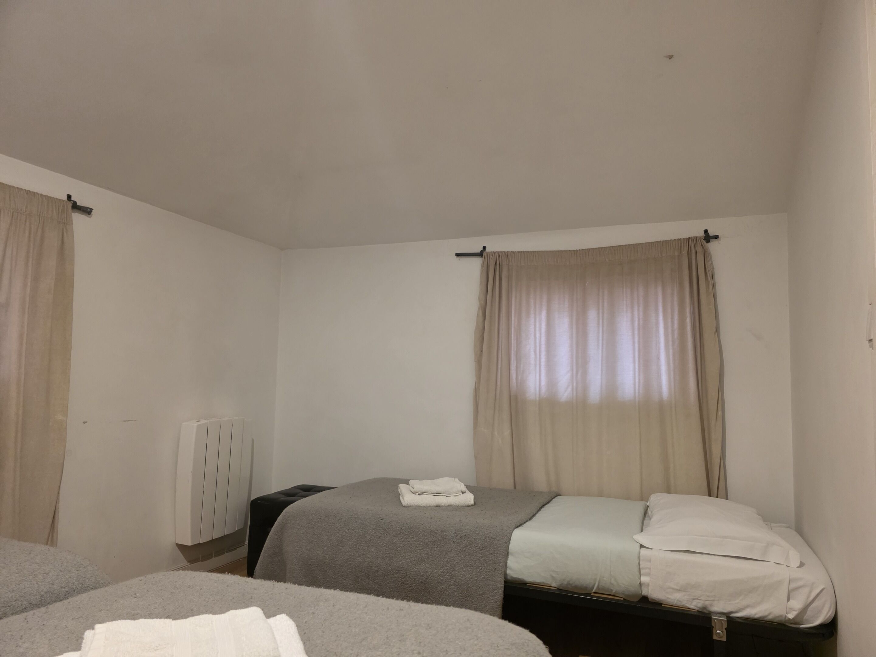 A room with two beds, beige curtains, a radiator, and a black ottoman.