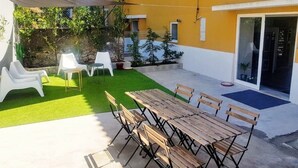 En udendørs patio med et træbord og stole, en hvid stol samt et lille haveområde med kunstgræs og krukkeplanter.