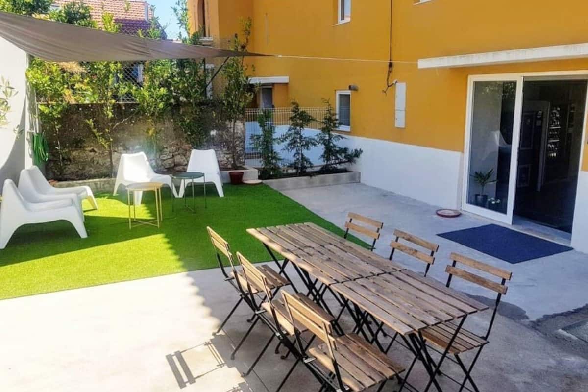 An outdoor patio with a wooden table and chairs, a white chair, and a small garden area with artificial grass and potted plants.