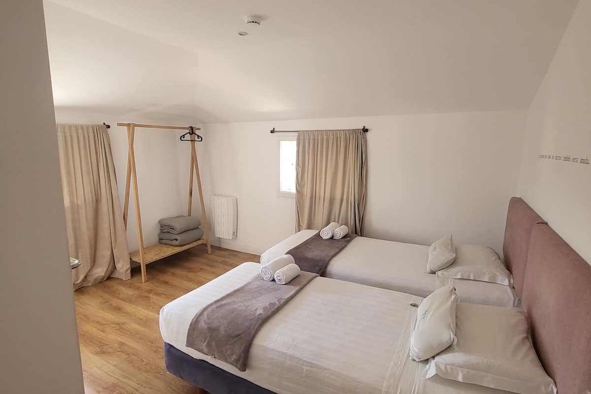 A bedroom with two beds, a wooden floor, a window with curtains, and a small table with a lamp.