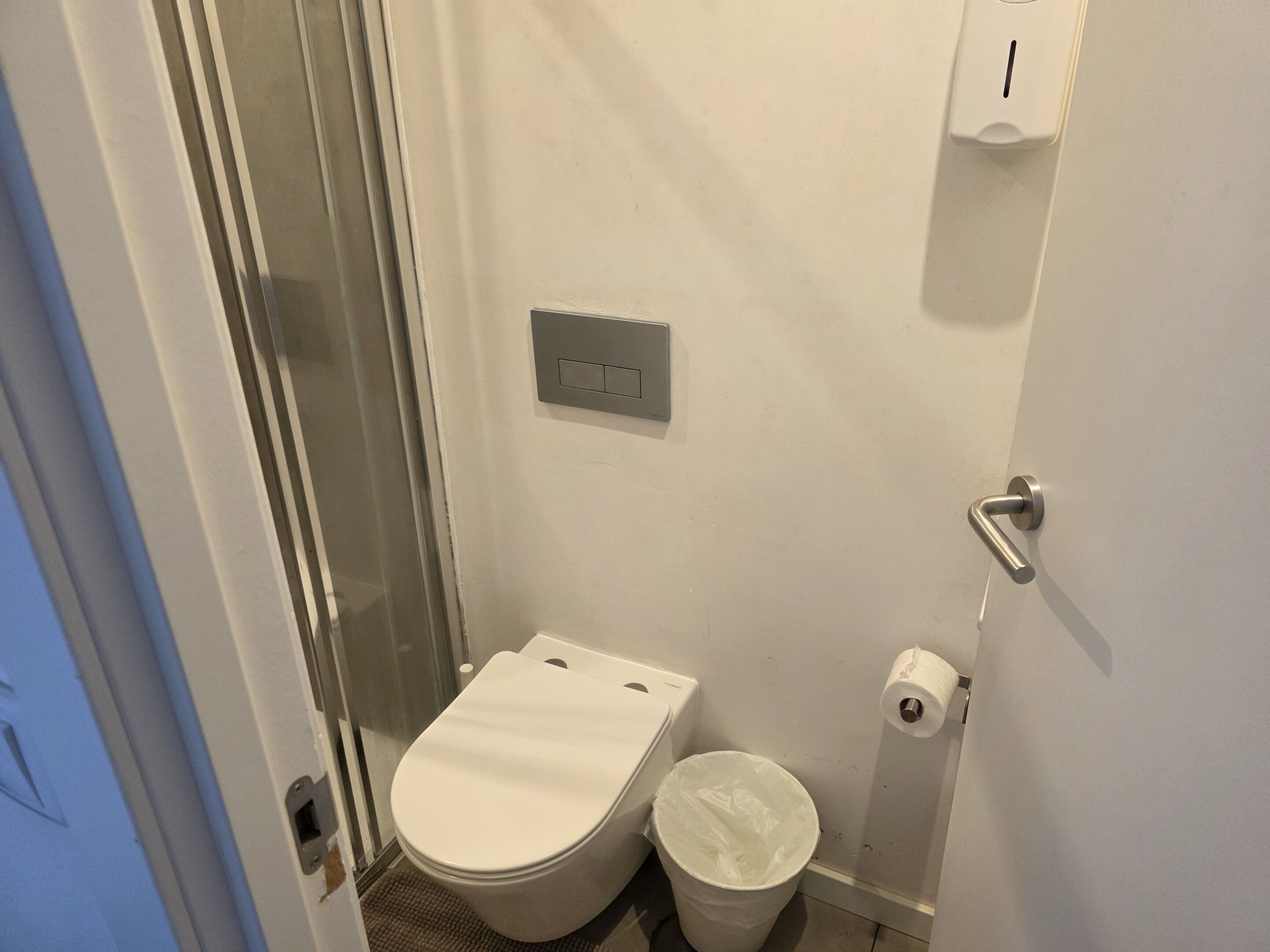 Une petite salle de bain compacte comprenant des toilettes, une poubelle, un distributeur d’essuie-tout et une cabine de douche en verre.