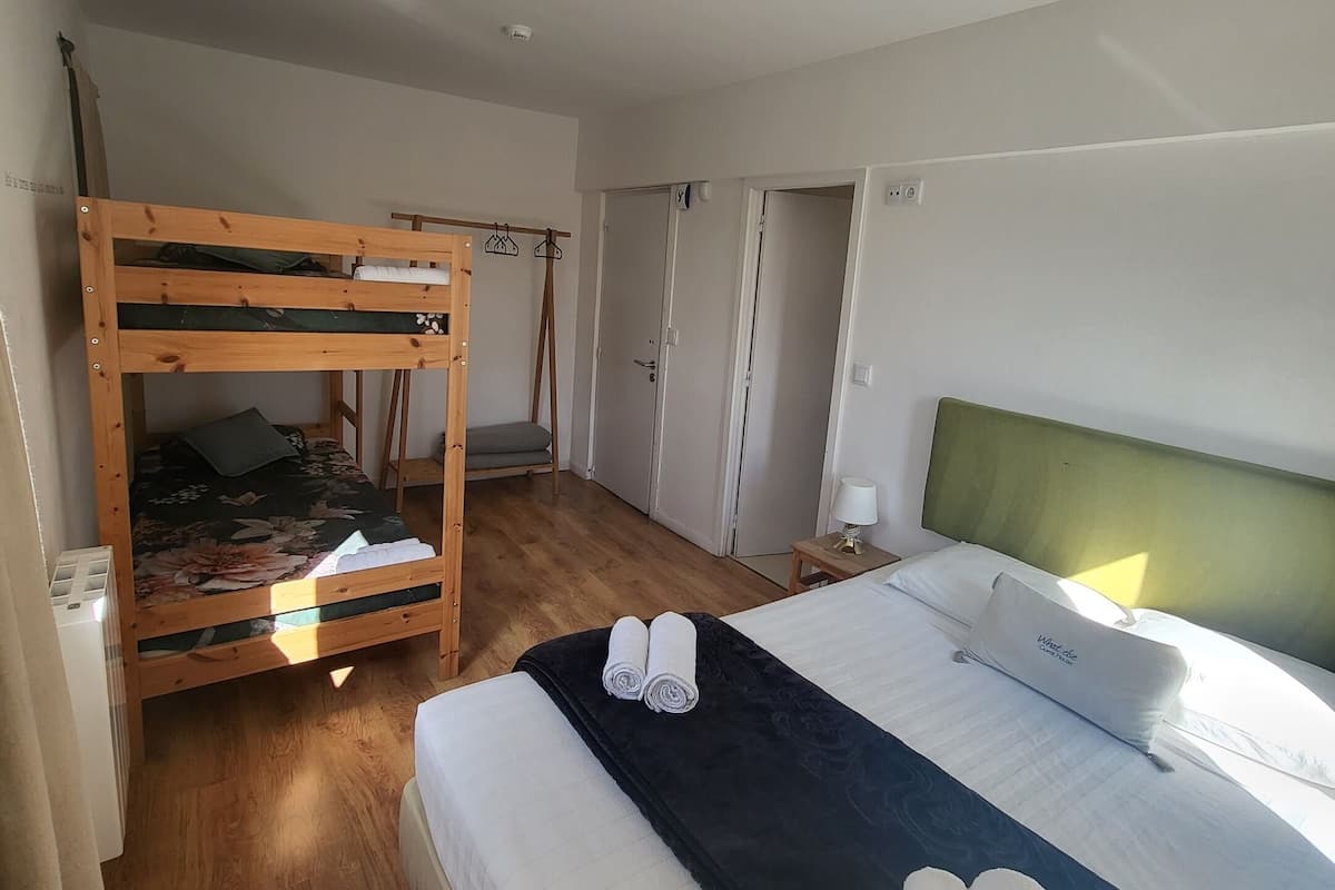 A bedroom with a bunk bed, a single bed, a bedside table with a lamp, and a window providing natural light.