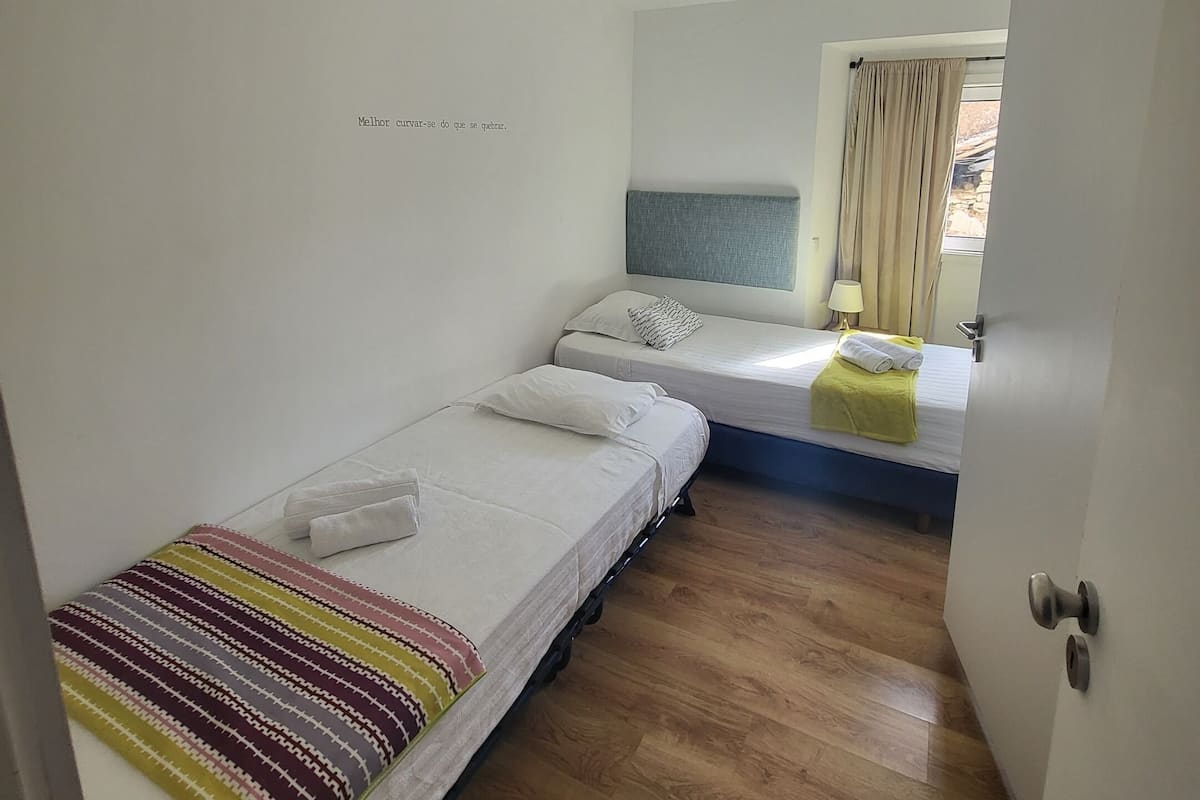 A small, clean room with two single beds, a window with curtains, and a wooden floor.