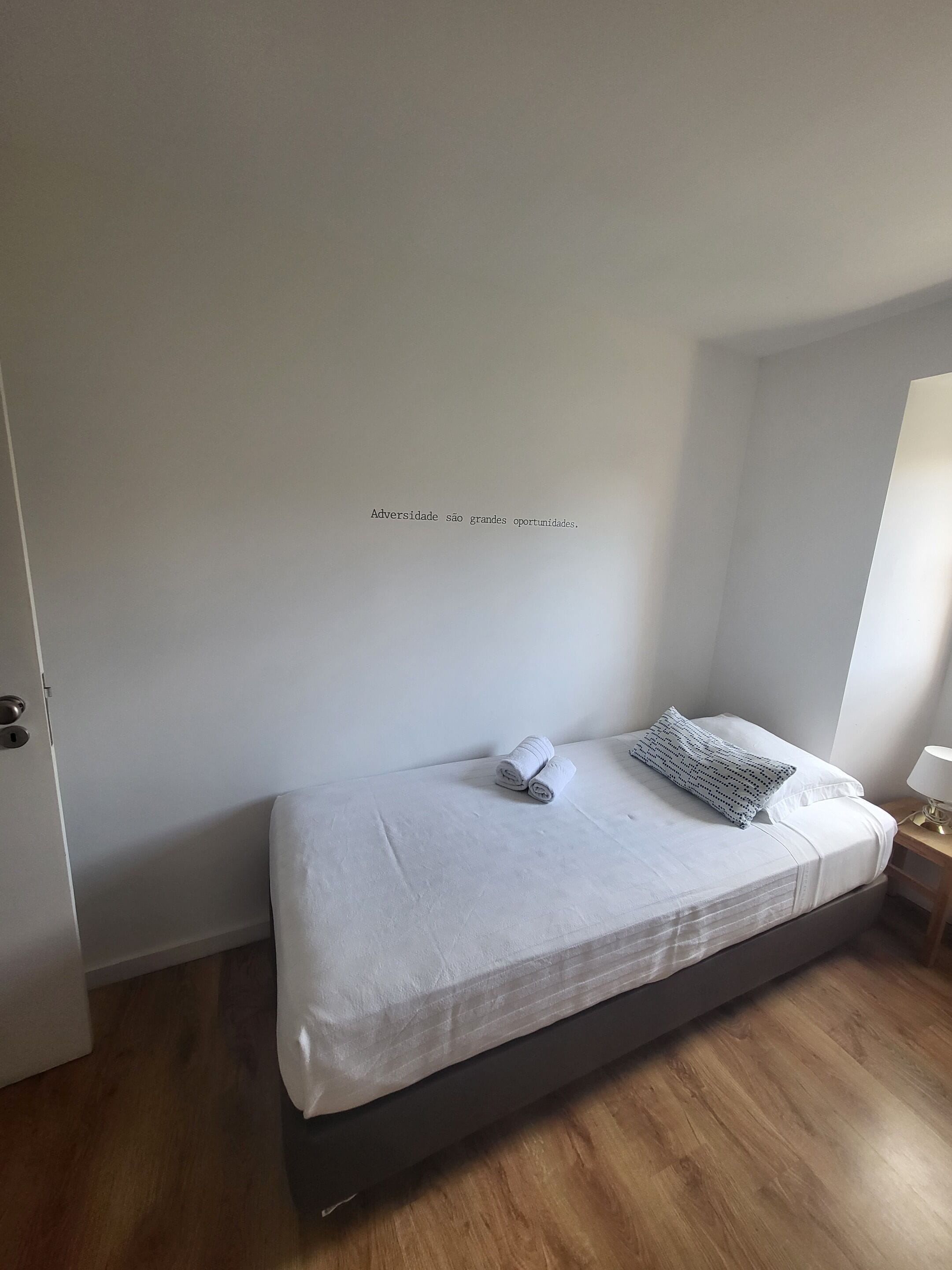 Una camera da letto minimalista con un letto singolo, un comodino con una lampada e una parete con una frase motivazionale.