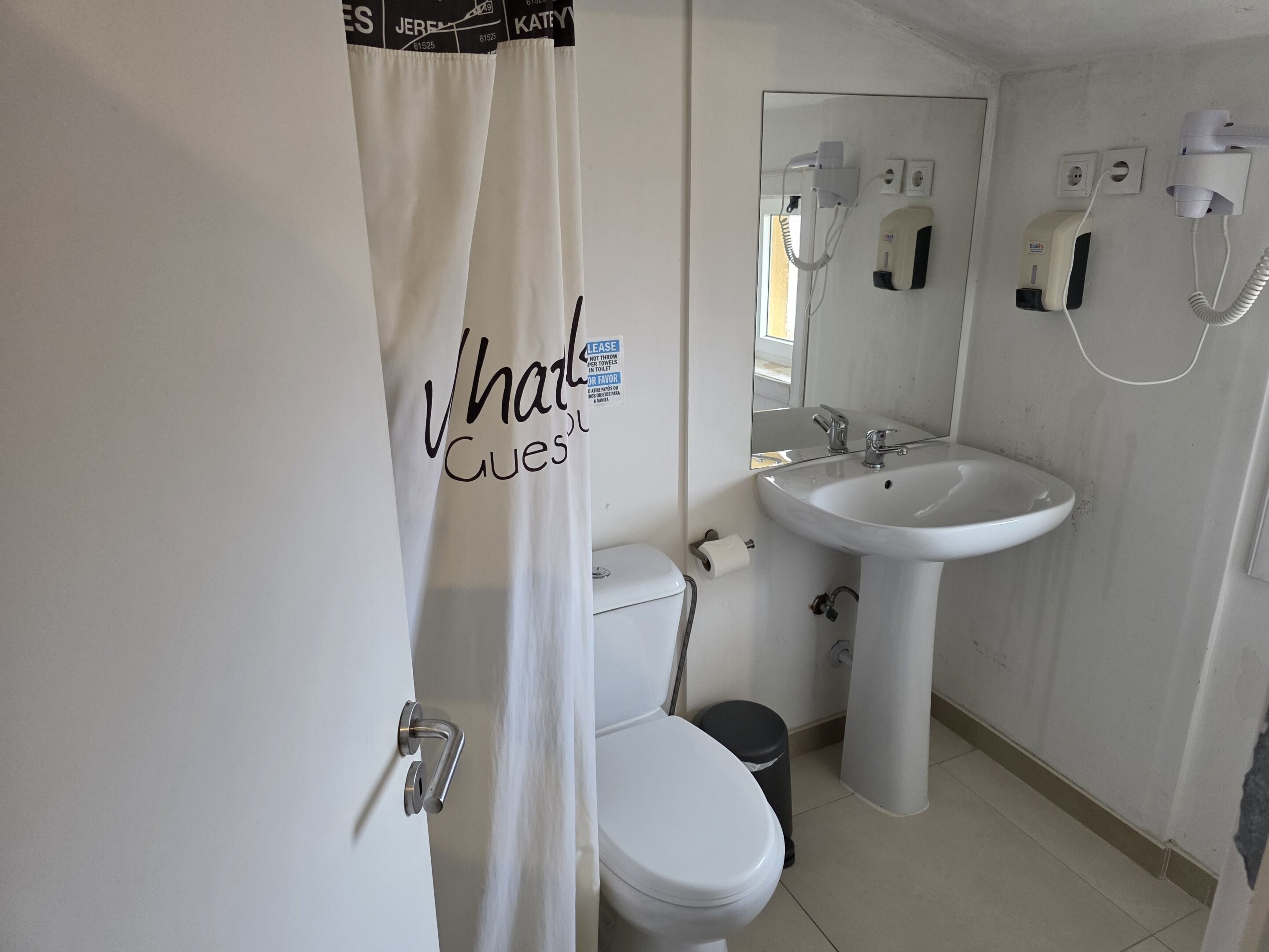 A bathroom with a white toilet, a sink, and a shower curtain. There is a mirror above the sink and a towel dispenser on the wall.