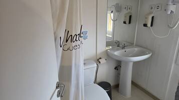 Une salle de bain avec des toilettes blanches, un lavabo et un rideau de douche. Il y a un miroir au-dessus du lavabo et un distributeur de serviettes au mur.
