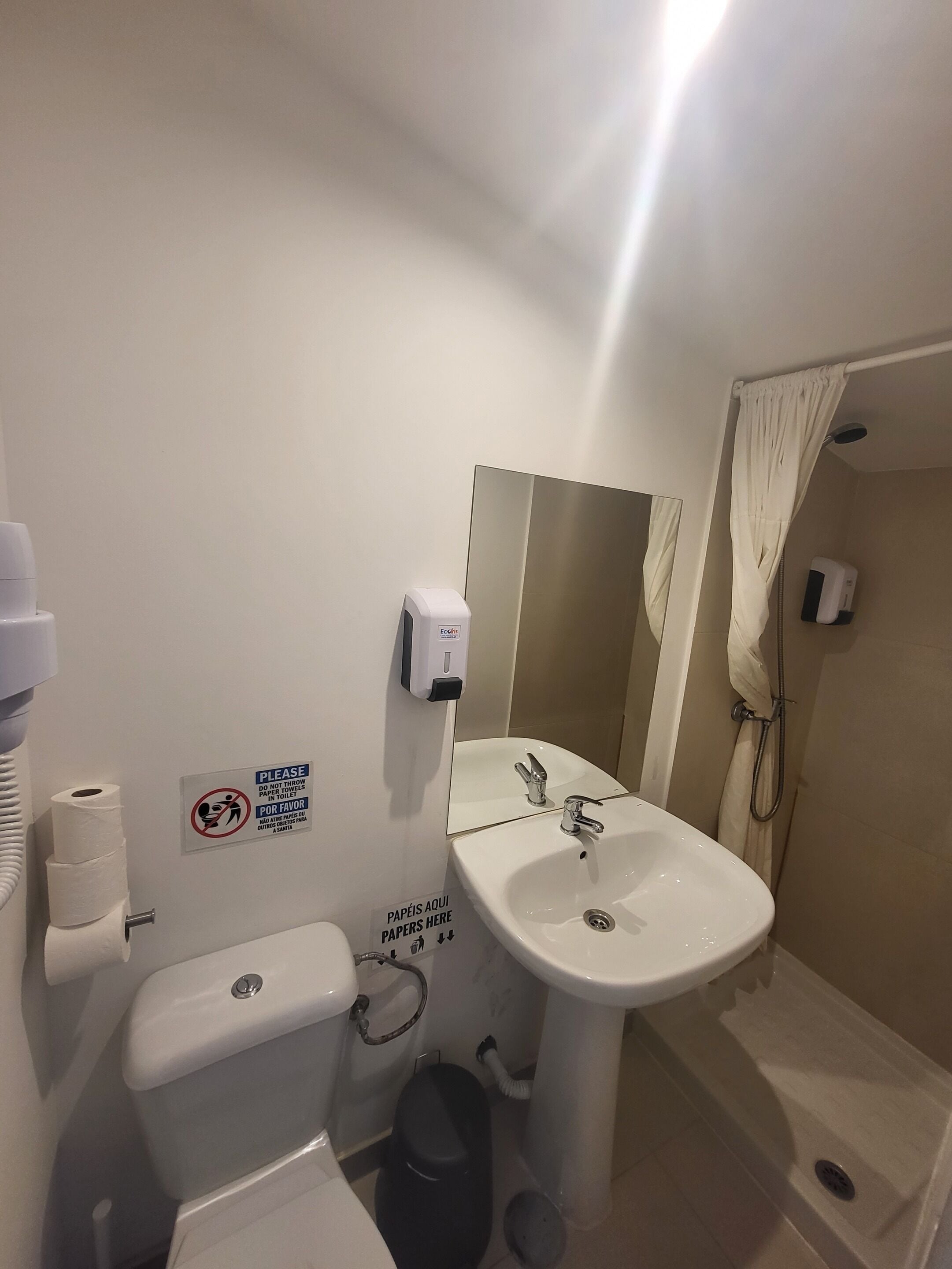 Un baño con inodoro, lavamanos y ducha. Hay letreros en la pared, un secador de manos y un cesto de basura.