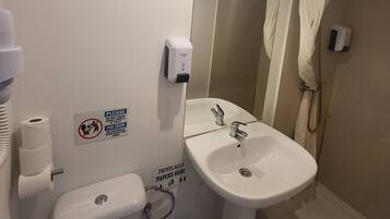 Un baño con inodoro, lavamanos y ducha. Hay letreros en la pared, un secador de manos y un cesto de basura.